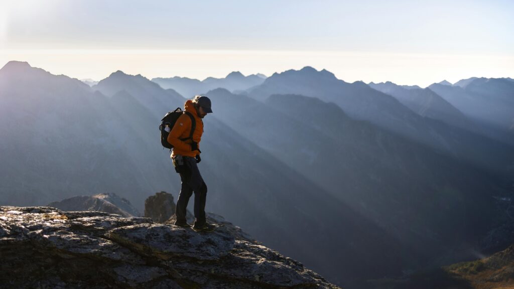 A lone hiker in an orange jacket explores a rocky peak at dawn, surrounded by breathtaking mountain views.